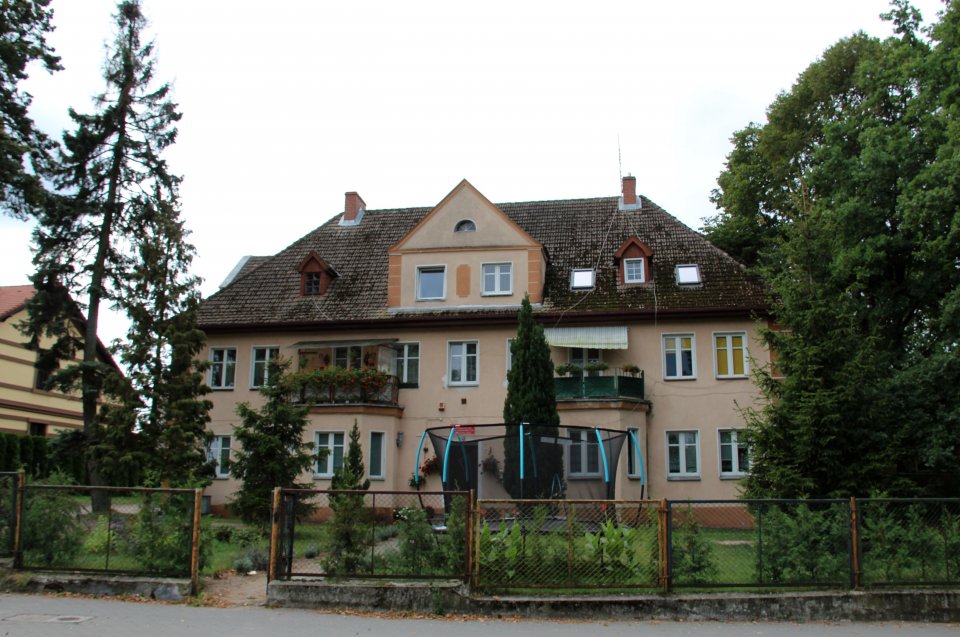 Budynek Placówki Opiekuńczo-Wychowawczej typu socjalizacyjnego w Jastrowiu. Przed nim krzewy oraz drzewa, a ogrodzie stoi trampolina. Z przodu ogrodzenie. Budynek Placówki Opiekuńczo-Wychowawczej typu socjalizacyjnego w Jastrowiu.